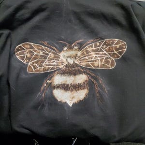 May include: Black hooded sweatshirt featuring a detailed illustration of a bee on the back. The bee has brown and white striped abdomen, brown wings with a webbed pattern, and a fuzzy head. The sweatshirt is made of a soft, dark fabric.