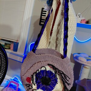 May include: A handmade, round, purple crochet bag with a large eye design. The eye features a blue iris, black pupil, and black eyelashes. The bag has a long strap with blue and black accents.