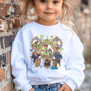 May include: A white sweatshirt featuring a colorful cartoon graphic. The graphic includes various animal characters in ornate frames, with a central green bow and floral accents. The child is wearing blue jeans.