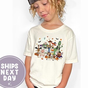 Puede incluir: Camiseta de manga corta color crema con un gráfico colorido con temática de Halloween de personajes animados. El diseño incluye calabazas, murciélagos y hojas de otoño. La camiseta la lleva un niño con una gorra de béisbol marrón.