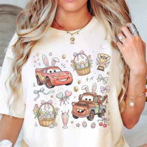Może przedstawiać: Kremowy t-shirt z kolorową grafiką o tematyce wielkanocnej. Projekt zawiera kresk&oacute;wkowe samochody z kr&oacute;liczymi uszami, koszyki wielkanocne, jajka i inne świąteczne elementy. Koszulka ma luźny kr&oacute;j.