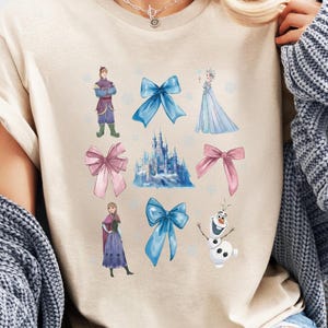 May include: Beige t-shirt featuring a colorful graphic of characters and bows from a popular animated movie. The design includes a castle, snowflakes, and various characters in shades of blue, pink, and purple. The shirt is made of a soft material.