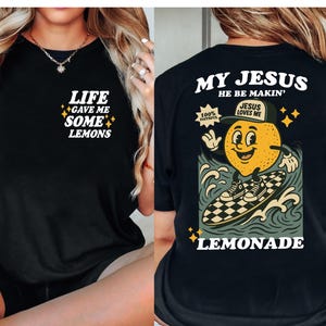 Puede incluir: Camiseta negra con el texto "LIFE GAVE ME SOME LEMONS" en la parte delantera. La parte trasera de la camiseta presenta un lim&oacute;n de dibujos animados surfeando en una tabla de surf a cuadros con el texto "MY JESUS HE BE MAKIN' LEMONADE".