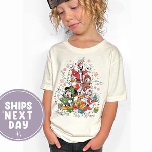Puede incluir: Camiseta de color crema con un gráfico navideño de personajes de Disney, incluyendo Mickey Mouse, Donald Duck y Goofy, con sus firmas. La camiseta tiene cuello redondo y mangas cortas. El niño lleva una gorra de béisbol marrón.