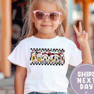 Puede incluir: Una camiseta blanca con un diseño festivo con personajes de Disney con gorros de Papá Noel y un borde a cuadros. Los personajes incluyen a Mickey Mouse, Donald Duck y Goofy. La niña lleva gafas de sol rosas y un lazo rosa.