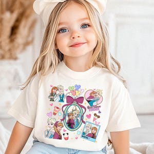 Puede incluir: Una niña pequeña con una camiseta crema con un gráfico colorido de personajes animados. El gráfico incluye el texto "Best Friends Forever" e imágenes de dos hermanas. La niña también lleva vaqueros azules y una diadema blanca.