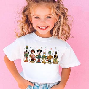 May include: A white t-shirt featuring a festive design of cartoon characters dressed as nutcrackers. The characters include Mickey Mouse, Minnie Mouse, Donald Duck, and others, with colorful accents and holiday lights. The shirt is worn by a young child.