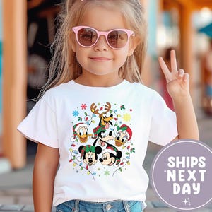 Puede incluir: Una camiseta blanca con un gráfico navideño de personajes de Disney, incluyendo Mickey Mouse, Donald Duck y Goofy, con gorros de Papá Noel y luces festivas. La niña lleva gafas de sol rosas y un lazo rosa. La imagen incluye el texto "SHIPS NEXT DAY".
