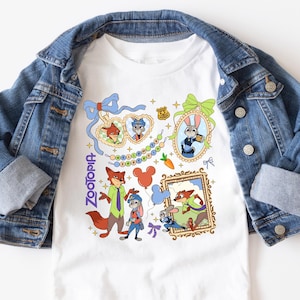 以下が含まれることがあります： 映画「ズートピア」のキャラクターが描かれた白いTシャツ。デザインには、額縁に入った肖像画、風船、そして縦書きの「Zootopia」の文字が含まれています。デニムジャケットがTシャツの上にかけられています。