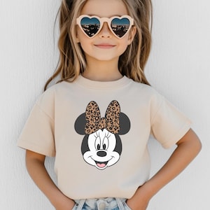 May include: A beige t-shirt featuring a Minnie Mouse graphic with a leopard print bow. The child is wearing heart-shaped sunglasses. The shirt is a short-sleeved crew neck.