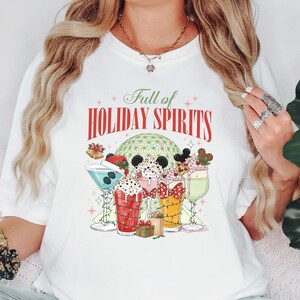 May include: White t-shirt with a colorful graphic featuring holiday-themed drinks. The design includes the text "Full of Holiday Spirits" above the drinks. The drinks are decorated with Mickey Mouse ears and other festive elements.