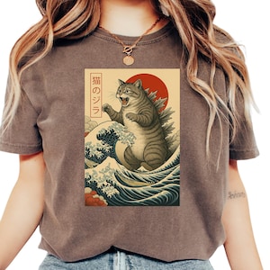 Könnte beinhalten: Braunes T-Shirt mit einem japanisch anmutenden Motiv einer katzenartigen Kreatur, einer roten Sonne und Meereswellen. Der japanische Text "猫のジラ" befindet sich auf der linken Seite des Motivs.