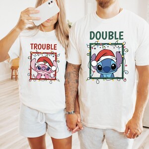 May include: Two white t-shirts with matching graphics. One shirt says "TROUBLE" and features a pink cartoon character wearing a Santa hat. The other says "DOUBLE" and has a blue cartoon character in a Santa hat.