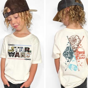 May include: Off-white Star Wars t-shirt with the Star Wars logo on the front and a graphic of characters on the back. The front logo features a colorful design. The back graphic includes Darth Vader, a Stormtrooper, and the Death Star.