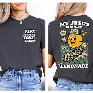 Puede incluir: Camiseta gris oscuro con el texto "LIFE GAVE ME SOME LEMONS" en la parte delantera. La parte trasera presenta un personaje de lim&oacute;n de dibujos animados surfeando en una tabla de surf a cuadros con las palabras "MY JESUS HE BE MAKIN' LEMONADE".