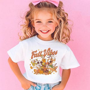 Op de afbeelding: Een wit T-shirt met de tekst "Fall Vibes" in oranje script. Het shirt heeft een cartoonafbeelding van Disney-personages. Het kind draagt een roze strik in haar haar en glimlacht.
