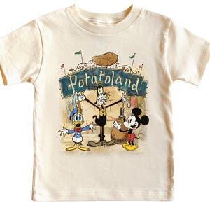 May include: Cream-colored toddler t-shirt featuring a colorful graphic of Disney characters Donald Duck, Goofy, and Mickey Mouse. The graphic includes the text "Potatoland" above the characters. The shirt has short sleeves and a crew neck.