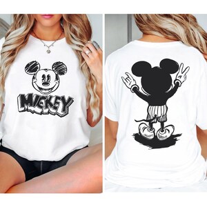 May include: White t-shirt featuring a black and white Mickey Mouse graphic on the front and back. The front graphic includes Mickey's face and the word "Mickey" in a bold, outlined font. The back graphic shows Mickey with his arms up in a rock and roll pose.