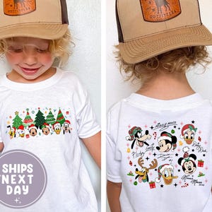 Puede incluir: Un niño lleva una camiseta blanca con un diseño navideño con personajes de Disney y árboles de Navidad. El niño también lleva una gorra de camionero marrón y beige con un logotipo. La camiseta tiene el texto "SHIPS NEXT DAY".