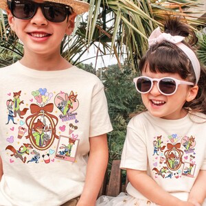 Może przedstawiać: Dwoje dzieci w kremowych koszulkach z kolorowym wzorem z kresk&oacute;wek. Wz&oacute;r przedstawia postać lisa w zielonej koszuli, kr&oacute;lika, balony oraz napisy "Best Friends Forever" i "Bestie".