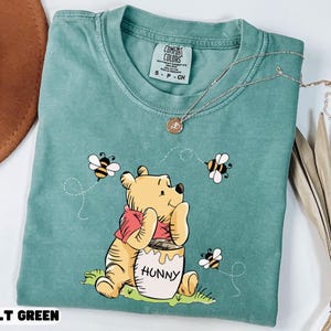 May include: A light green t-shirt featuring a cartoon bear with a red scarf, sitting next to a honey pot labeled "Hunny". The shirt also has three bees and the text "LT GREEN". A silver necklace with a pendant is on top of the shirt.