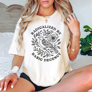 Könnte beinhalten: Cremefarbenes T-Shirt mit einer schwarzen Grafik eines Vogels, umgeben von Blättern und Blumen, und dem Text "RADICALIZED BY BASIC DECENCY". Das Shirt hat kurze Ärmel und eine lockere Passform.