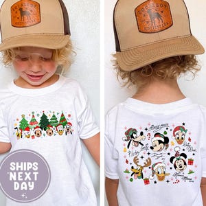 Puede incluir: Un niño lleva una camiseta blanca con un diseño navideño con personajes de Disney y árboles de Navidad. El niño también lleva una gorra de camionero marrón y beige con un logotipo. La camiseta tiene el texto "SHIPS NEXT DAY".