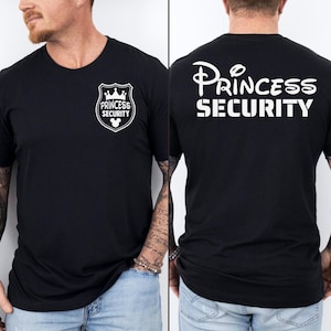 Puede incluir: Camiseta negra de cuello redondo con texto y gráficos blancos. La parte delantera presenta un diseño de escudo con las palabras "PRINCESS SECURITY" y una corona y la silueta de Mickey Mouse. La parte trasera de la camiseta muestra las palabras "PRINCESS SECURITY".