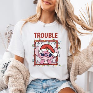 May include: White t-shirt featuring a pink cartoon character wearing a Santa hat, surrounded by Christmas lights, and the word "TROUBLE" in red. The design is enclosed in a red square.
