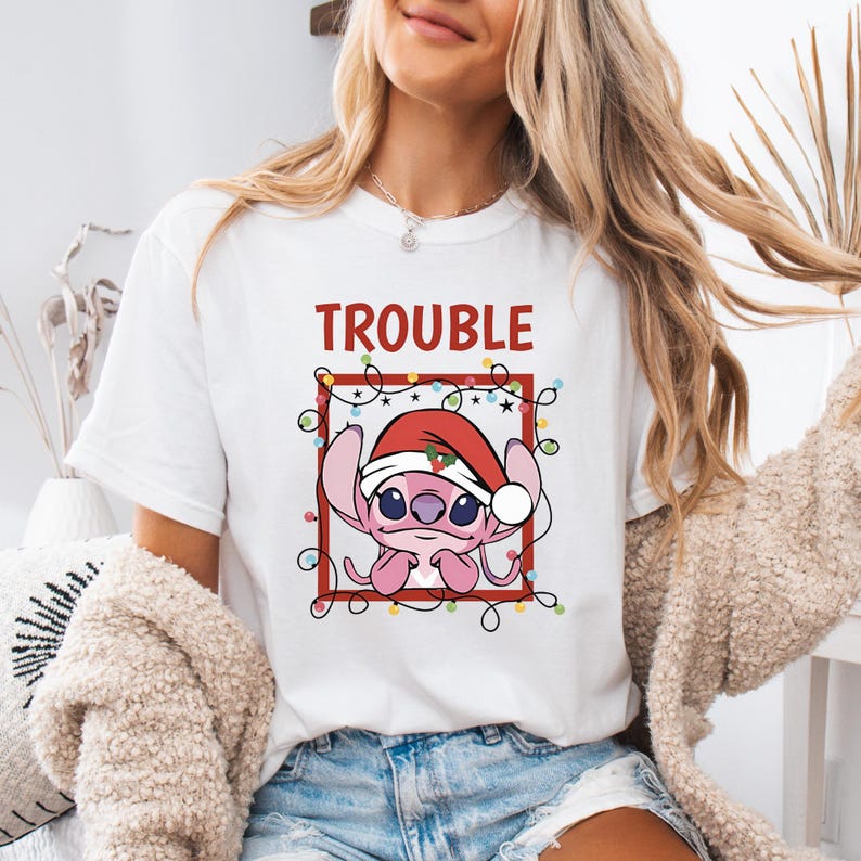 May include: White t-shirt featuring a pink cartoon character wearing a Santa hat, surrounded by Christmas lights, and the word "TROUBLE" in red. The design is enclosed in a red square.