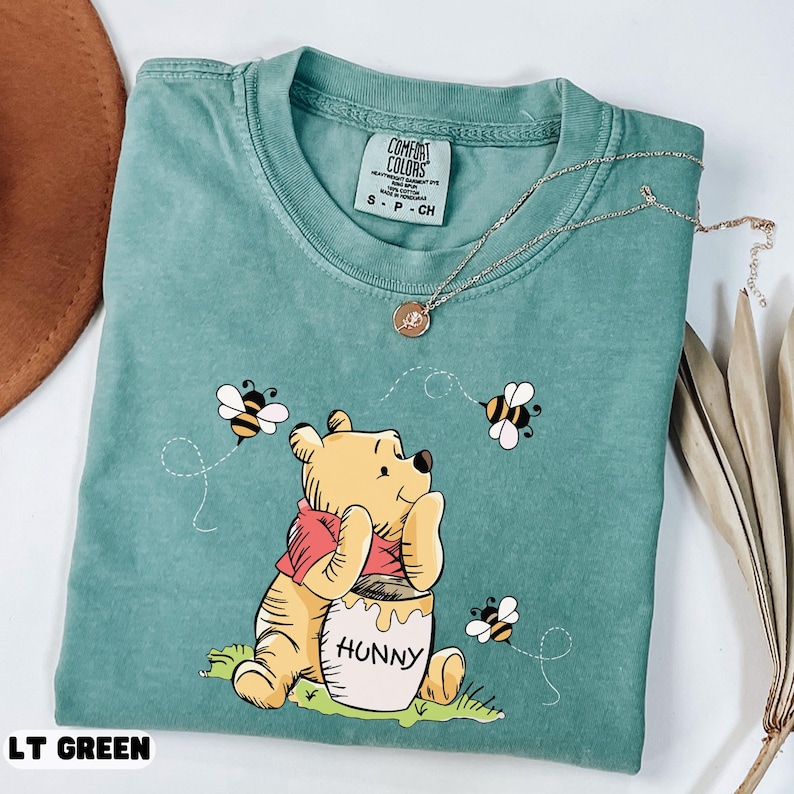 May include: A light green t-shirt featuring a cartoon bear with a red scarf, sitting next to a honey pot labeled "Hunny". The shirt also has three bees and the text "LT GREEN". A silver necklace with a pendant is on top of the shirt.