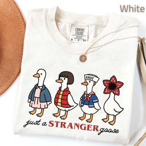 May include: White t-shirt with a graphic of four cartoon geese dressed as characters from a popular TV show. The text "just a STRANGER goose" is printed below the geese. A gold necklace is draped across the shirt.