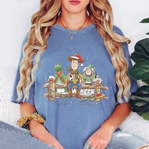 Puede incluir: Camiseta azul con una ilustraci&oacute;n festiva de Woody, Rex y Buzz Lightyear de Toy Story, adornada con decoraciones navide&ntilde;as. El dise&ntilde;o incluye una escena nevada y una valla de madera.