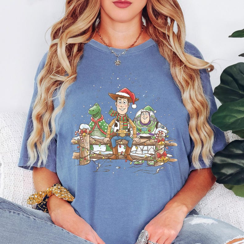 Puede incluir: Camiseta azul con una ilustraci&oacute;n festiva de Woody, Rex y Buzz Lightyear de Toy Story, adornada con decoraciones navide&ntilde;as. El dise&ntilde;o incluye una escena nevada y una valla de madera.