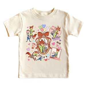 Może przedstawiać: Kremowa koszulka z postaciami z kresk&oacute;wki Zootopia. Projekt zawiera Nicka Wilde'a i Judy Hopps, a także tekst "Best Friends Forever" i "Bestie". Grafika zawiera r&oacute;wnież serca, rożki lod&oacute;w i balony.