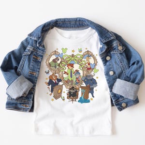 May include: White t-shirt featuring a colorful cartoon graphic of various animal characters in a heart-shaped frame. The design includes a fox, bunny, lion, and other animals. A denim jacket is draped over the shirt.