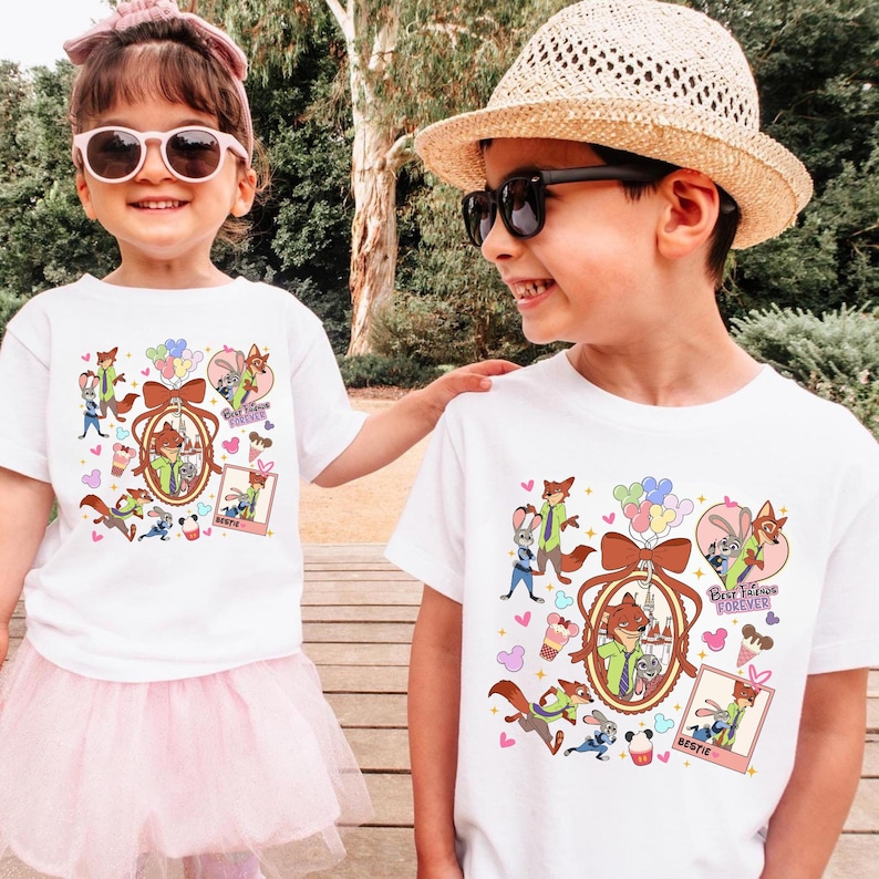 Może przedstawiać: Dwoje dzieci w białych koszulkach z kolorowym wzorem z kresk&oacute;wek. Wz&oacute;r zawiera postacie z kresk&oacute;wek, balony oraz napisy "Best Friends Forever" i "Bestie". Dziewczynka ma na sobie r&oacute;żową tutu i okulary przeciwsłoneczne.