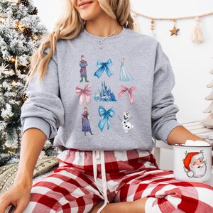 May include: A gray sweatshirt featuring watercolor illustrations of characters, bows, and a castle. The design includes a blue castle, pink and blue bows, and characters in purple, blue, and green. The sweatshirt is part of a winter holiday theme.