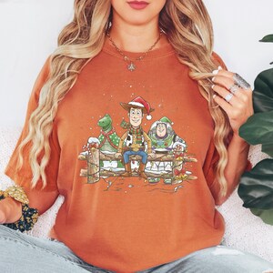 Puede incluir: Una camiseta naranja con un dise&ntilde;o navide&ntilde;o. Presenta a los personajes de dibujos animados Woody, Rex y Buzz Lightyear, sentados en una valla de madera decorada con luces y nieve, con sombreros y bufandas festivas.