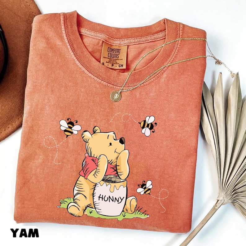 May include: Orange Comfort Colors long-sleeve shirt featuring a cartoon illustration of Winnie the Pooh with a honey pot and bees. A gold necklace and the word "YAM" are also visible.