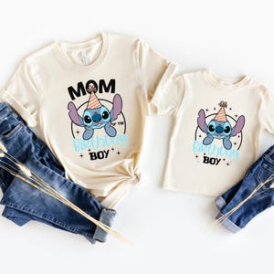 K&ouml;nnte beinhalten: Zwei cremefarbene T-Shirts mit einem Cartoon-Charakter-Design. Auf einem Shirt steht "MOM OF THE BIRTHDAY BOY" und auf dem anderen "BIRTHDAY BOY". Beide Shirts haben eine blaue Cartoon-Figur mit Partyhut.