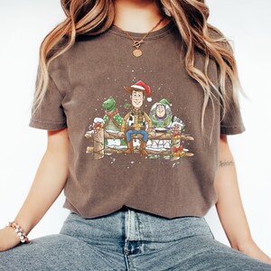 Puede incluir: Camiseta marr&oacute;n con una ilustraci&oacute;n navide&ntilde;a de Woody, Rex y Buzz Lightyear. Woody lleva un gorro de Pap&aacute; Noel y est&aacute; sentado en una valla de madera con los otros personajes. El dise&ntilde;o incluye nieve y decoraciones.