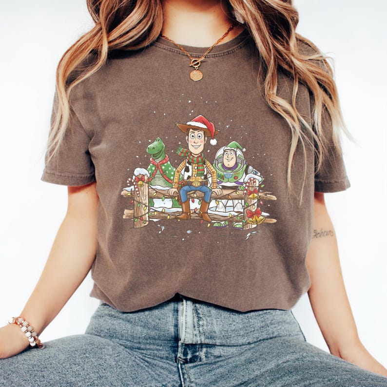 Puede incluir: Camiseta marr&oacute;n con una ilustraci&oacute;n navide&ntilde;a de Woody, Rex y Buzz Lightyear. Woody lleva un gorro de Pap&aacute; Noel y est&aacute; sentado en una valla de madera con los otros personajes. El dise&ntilde;o incluye nieve y decoraciones.