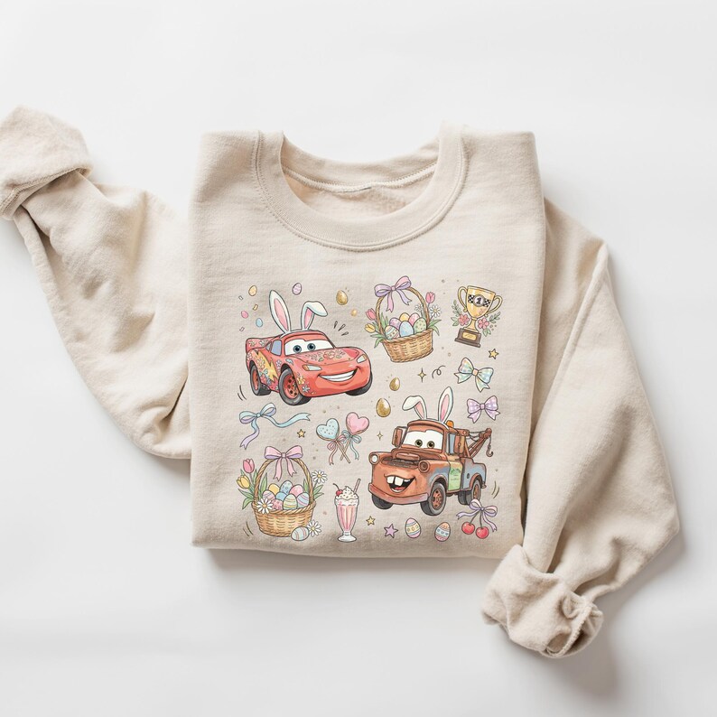 Może przedstawiać: Beżowa bluza z kresk&oacute;wkowymi ilustracjami Zygzaka McQueena i Złomka z Aut, obaj z kr&oacute;liczymi uszami. Grafika w tematyce wielkanocnej obejmuje kosze z jajkami, kokardy, trofeum i koktajl mleczny. Projekt jest w miękkich pastelowych kolorach.