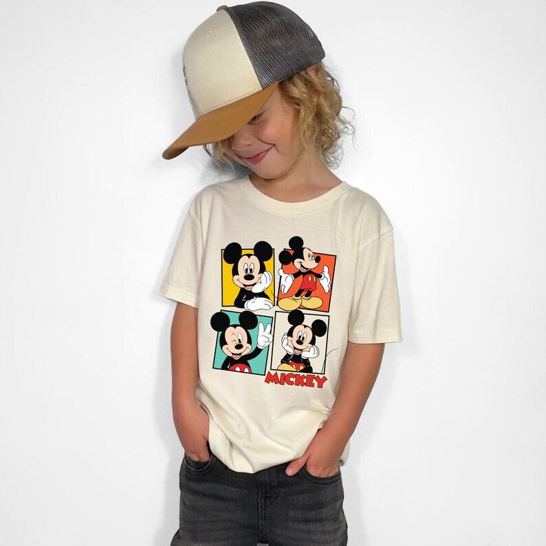May include: A child wearing a cream-colored t-shirt featuring four panels with different Mickey Mouse poses. The word "Mickey" is printed in red. The child is also wearing a corduroy baseball cap with a brown brim.