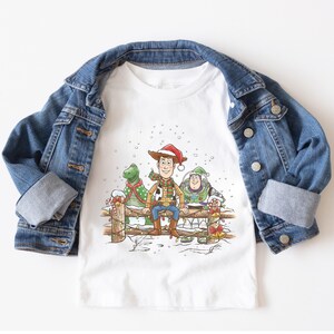 Puede incluir: Camiseta blanca con una ilustraci&oacute;n festiva de Woody, Rex y Buzz Lightyear de Toy Story, sentados en una valla de madera en una escena nevada. Los personajes est&aacute;n adornados con decoraciones navide&ntilde;as, y la camiseta se combina con una chaqueta vaquera.