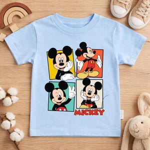 May include: A light blue t-shirt featuring four panels with different poses of Mickey Mouse. The word "MICKEY" is printed in red below the panels. The shirt is made of a soft material and is suitable for children.