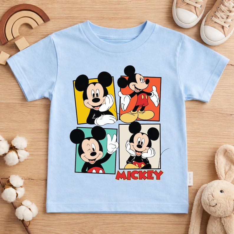 May include: A light blue t-shirt featuring four panels with different poses of Mickey Mouse. The word "MICKEY" is printed in red below the panels. The shirt is made of a soft material and is suitable for children.