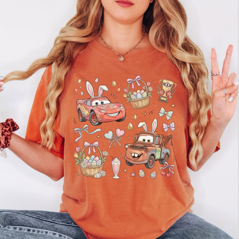 Może przedstawiać: Pomarańczowa koszulka z nadrukiem w kolorze spalonej pomarańczy z kolorową grafiką wielkanocną. Projekt zawiera kresk&oacute;wkowe samochody z kr&oacute;liczymi uszami, koszyki wielkanocne, jajka, kokardy i trofeum. Koszulka jest wygodna i swobodna.