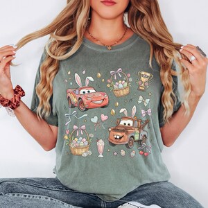 Może przedstawiać: Szałwiowa zielona koszulka z wielkanocnym wzorem. Grafika przedstawia kresk&oacute;wkowe samochody z kr&oacute;liczymi uszami, pisanki i kosze kwiat&oacute;w. Projekt przedstawia czerwony samoch&oacute;d, brązową lawetę i pastelowe kokardy. Koszulka jest w stylu casual.